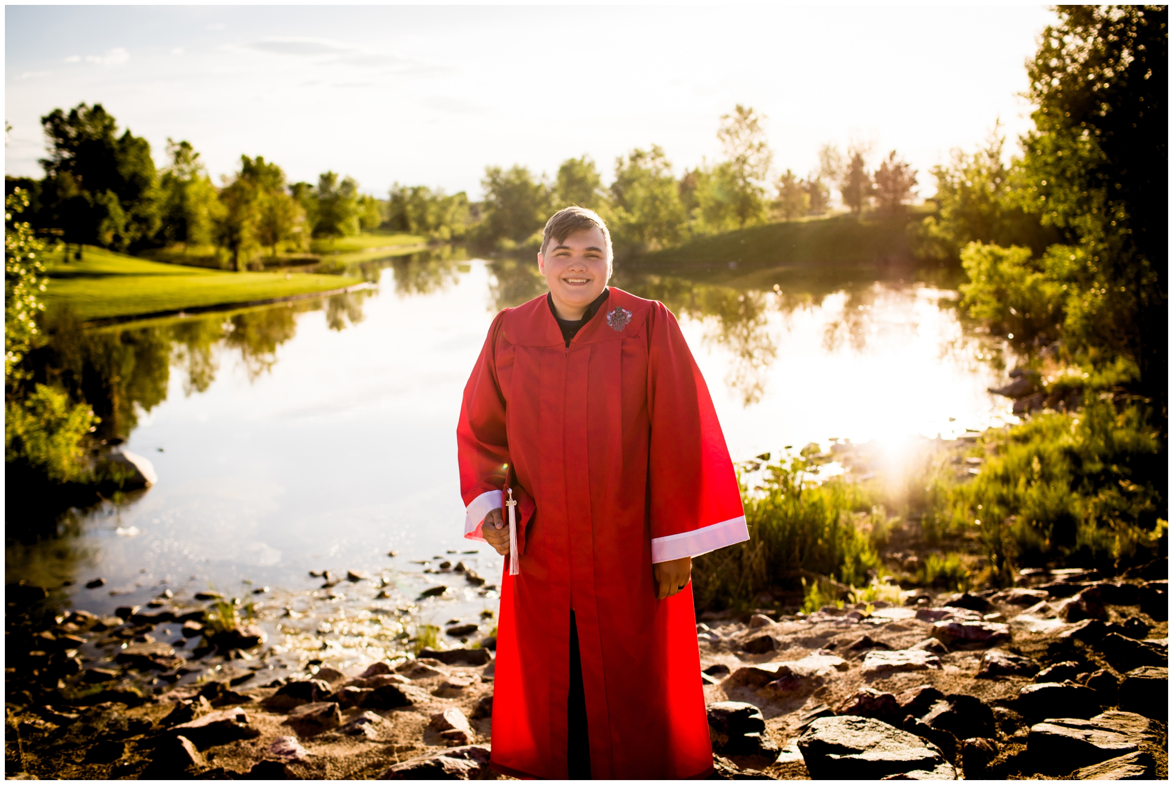 teen boy in cap and gown posing in front of lake during Brighton High School Colorado senior pictures 