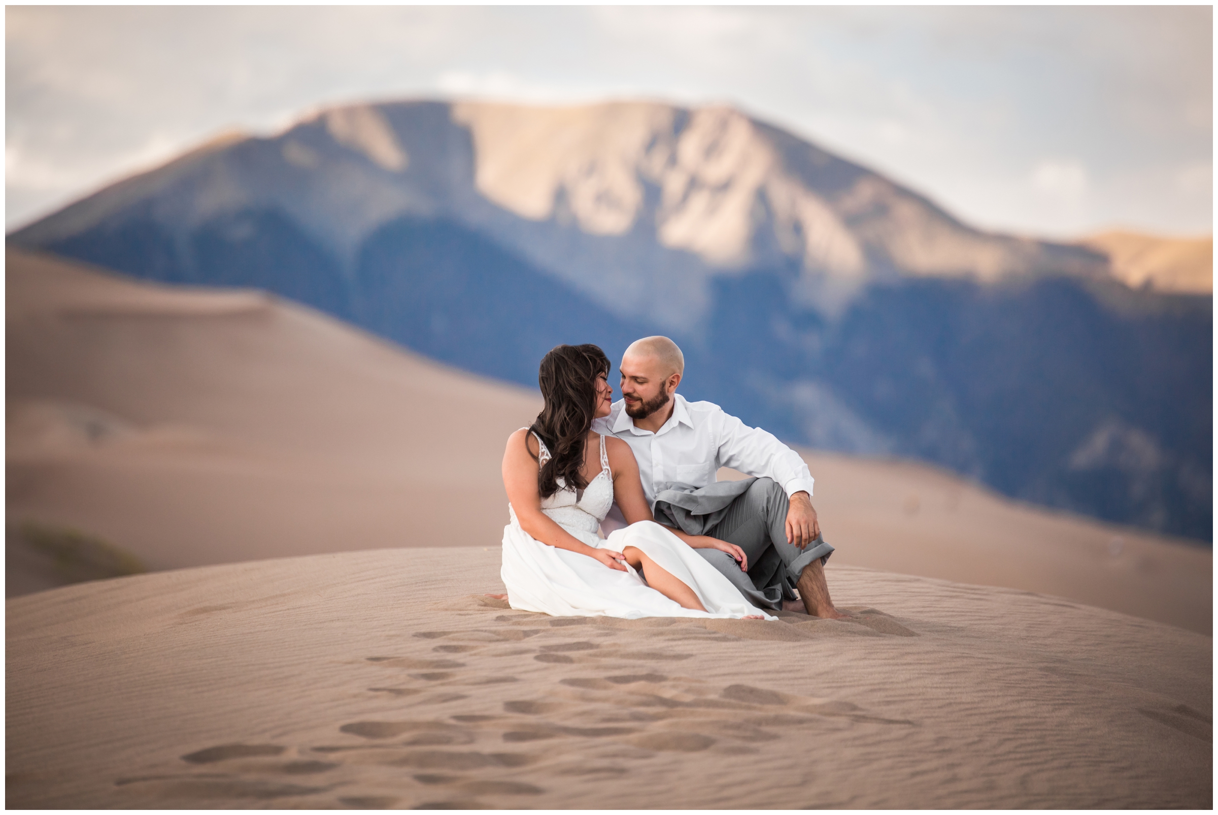couple sitting in the sand with mountains in background during Great Sand Dunes Colorado engagement portraits 