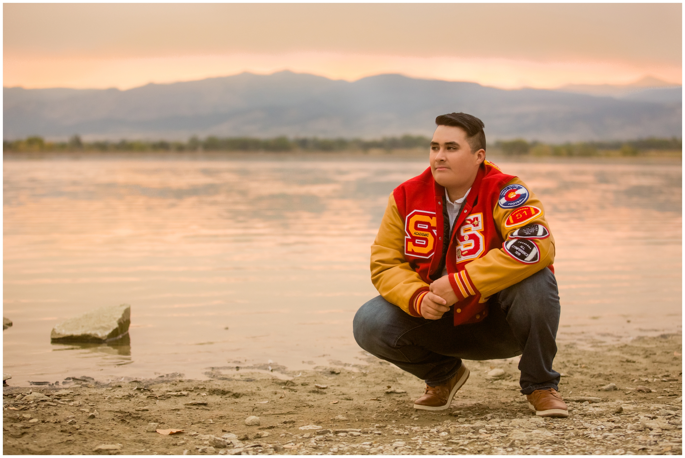 teen boy in letterman jacket posing next to lake at sunset at Longmont graduation photography session 