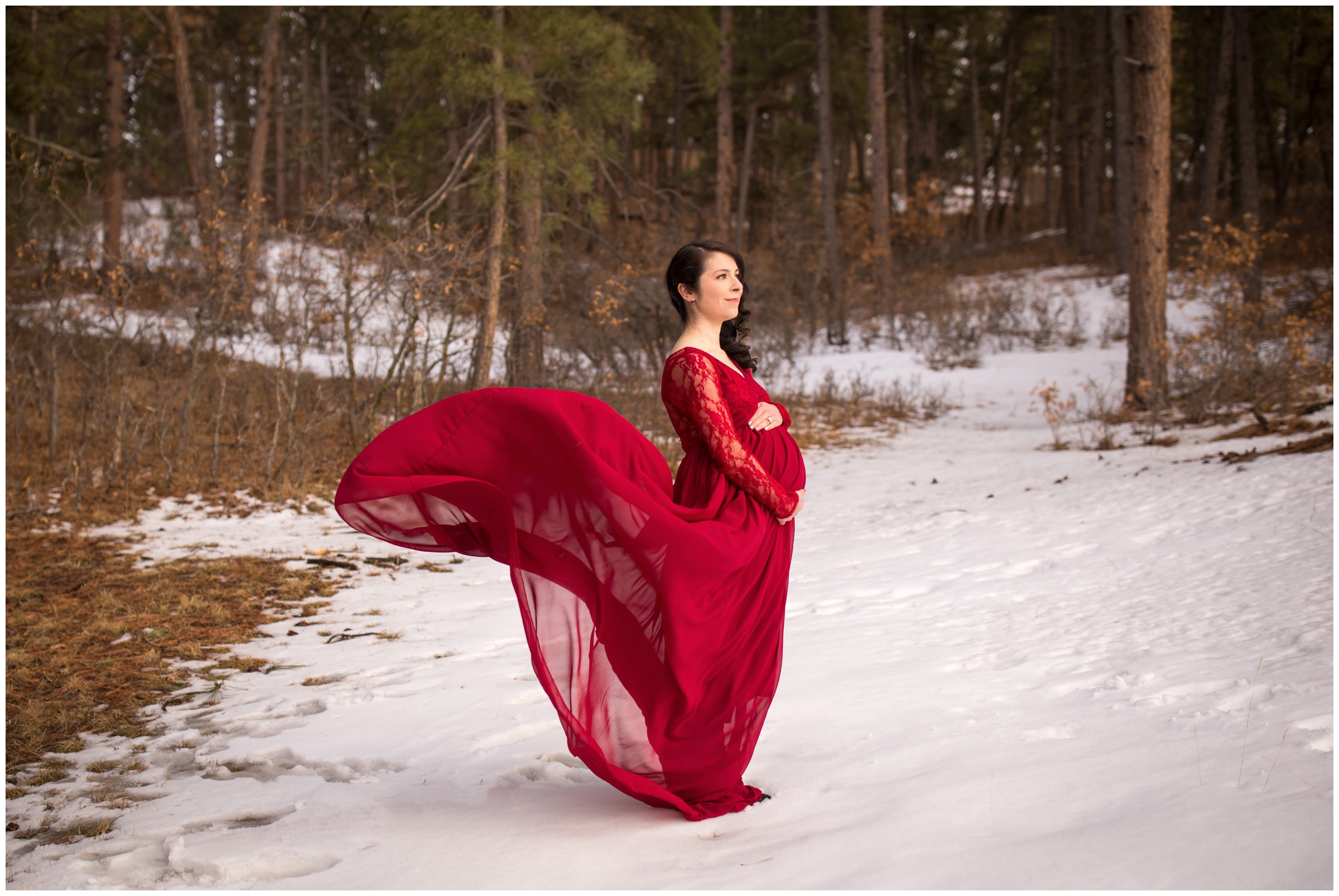 woman in red dress standing in the snow during Colorado winter maternity photos in a forest setting
