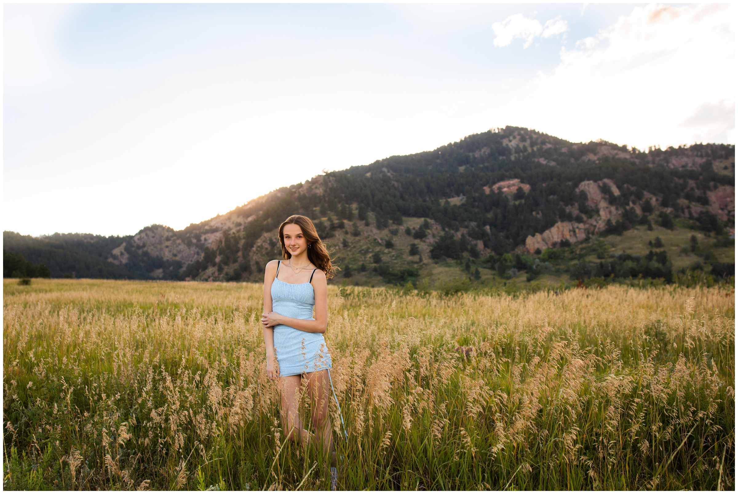 teen in light blue dress posing in golden field with mountains in background during Colorado senior portraits 