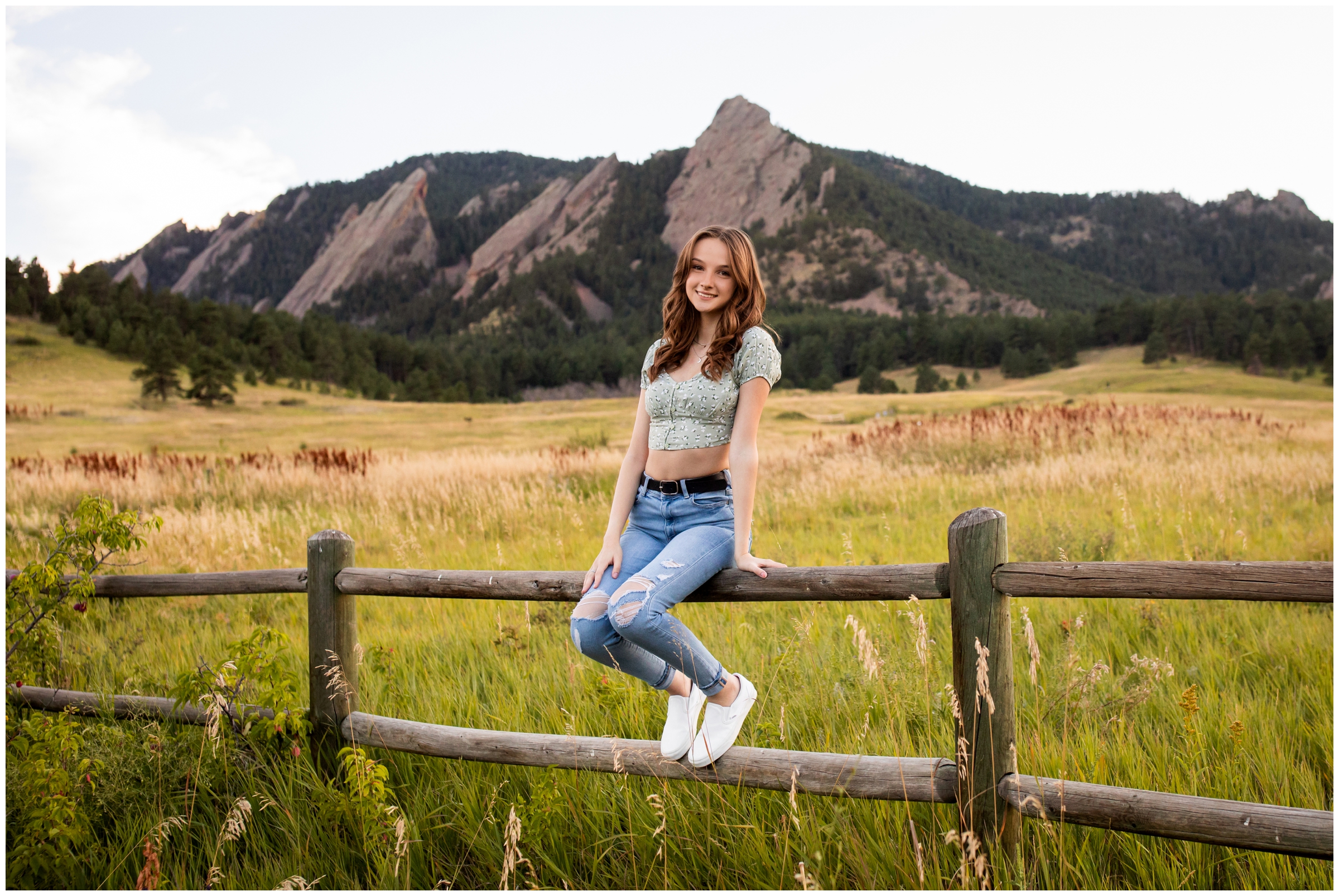 teen girl sitting on wooden fence with flatirons in background during Chautauqua Park Colorado senior portraits
