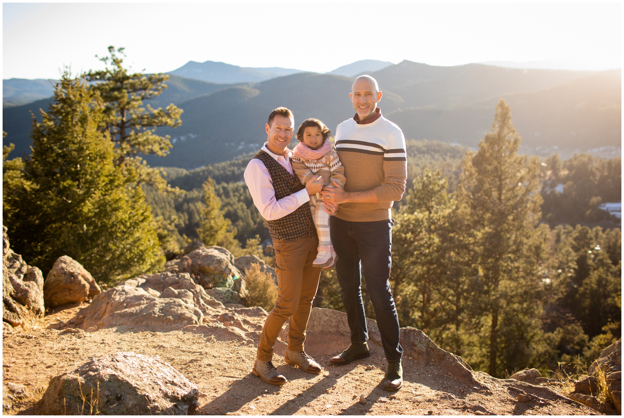 two dads holding daughter with mountains in background during Colorado Mount Falcon family photography session 