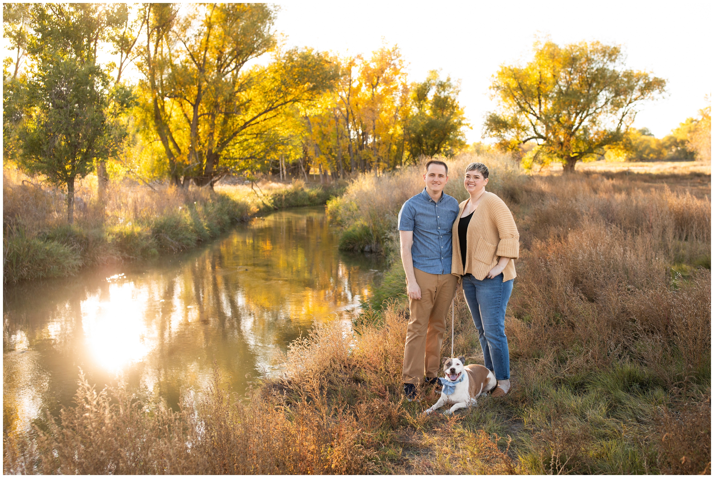 couple and their dog posing next to a river during colorful fall photography session at Sandstone Ranch in Longmont