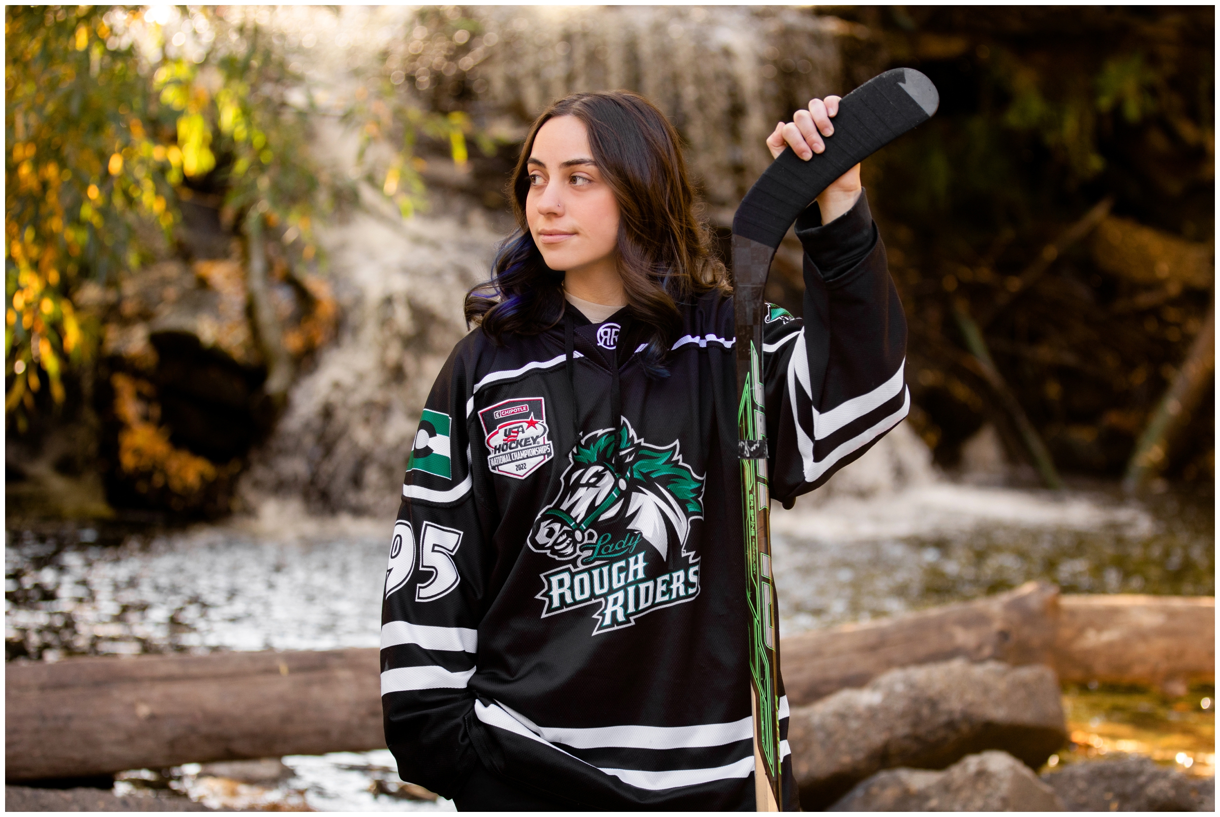 teen posing in front of waterfall with hockey stick in hand during Colorado hockey senior pictures in Longmont
