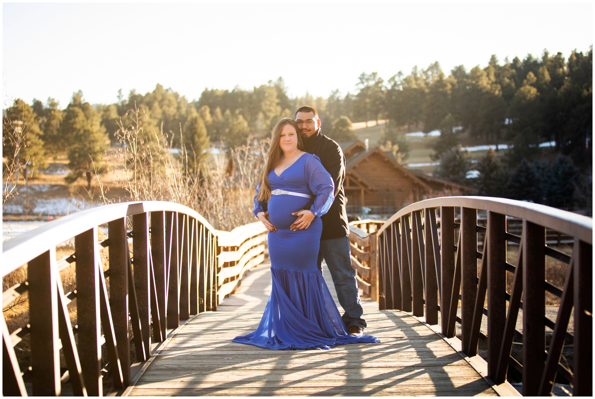 couple posing on bridge with Evergreen Lake House in background during winter maternity pictures in Colorado