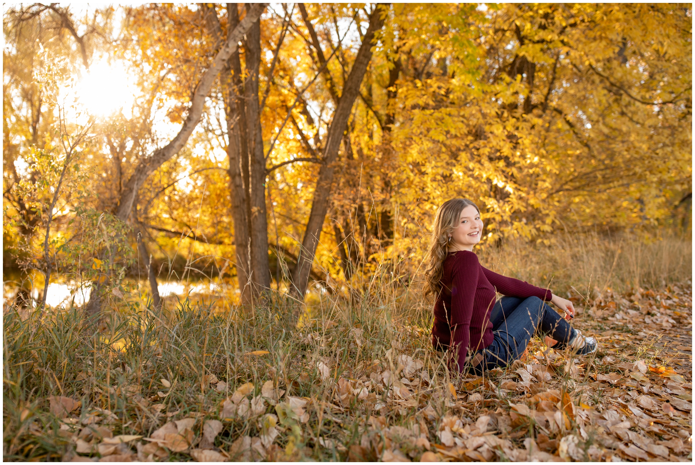 teen sitting in pile of crunchy leaves with fall foliage in background during senior portraits in Longmont Colorado