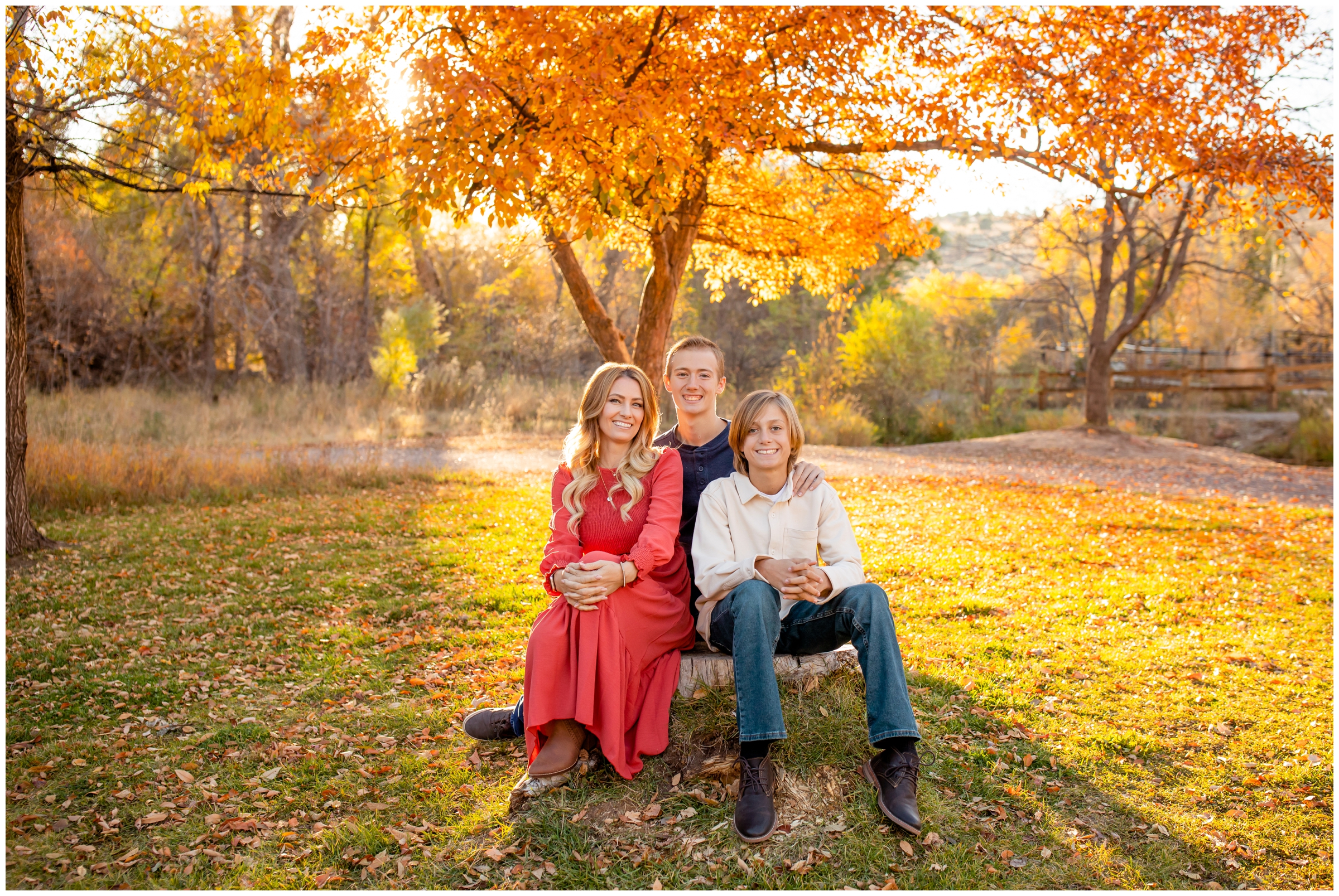 adult sons and mom sitting on large rock with fall foliage in background during Lyons Colorado family photography session at Bohn Park