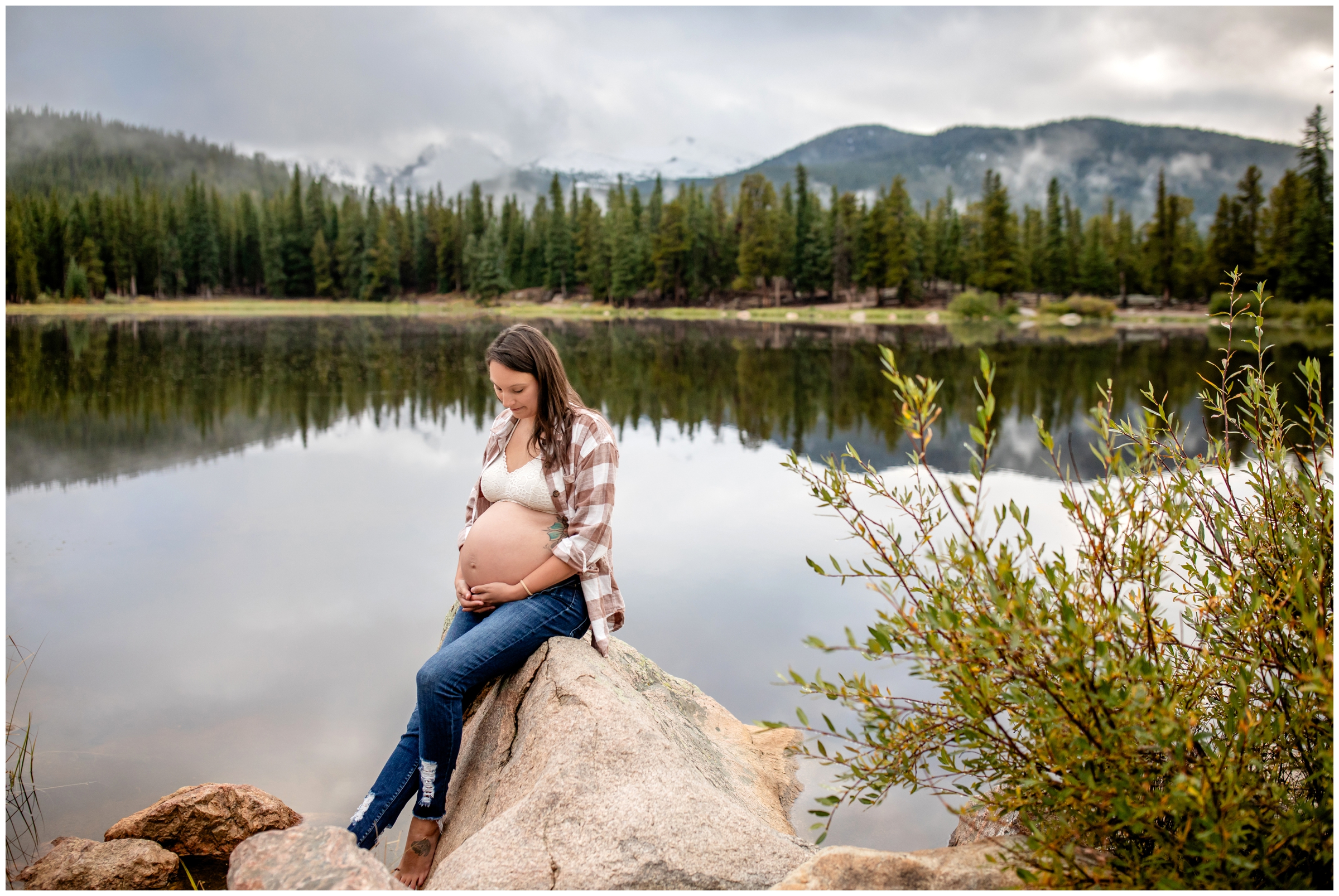 pregnant woman sitting on a rock with mountains and lake in background during Colorado maternity portraits by Plum Pretty Photography