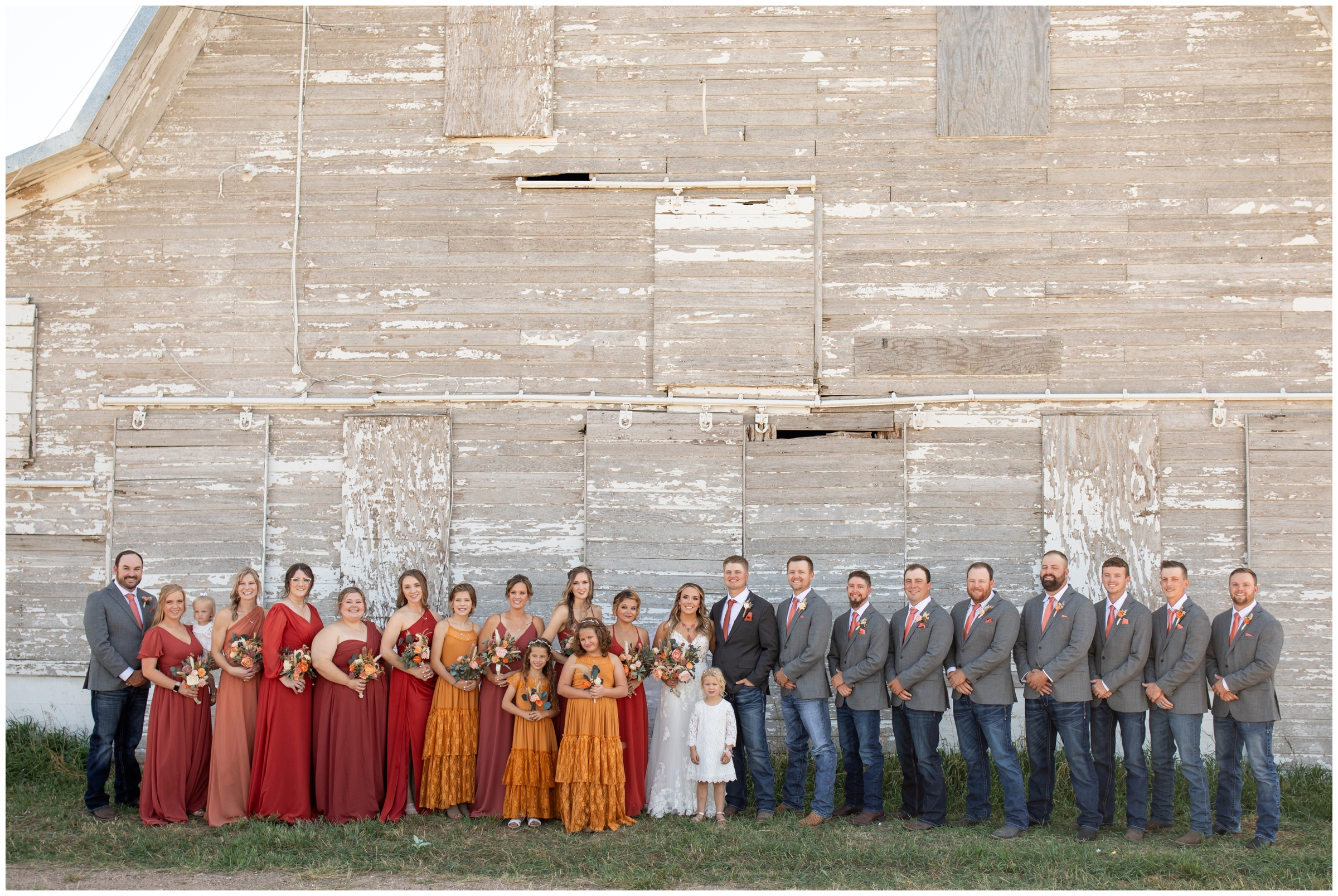 large wedding party in shades of red and orange  in front of barn 