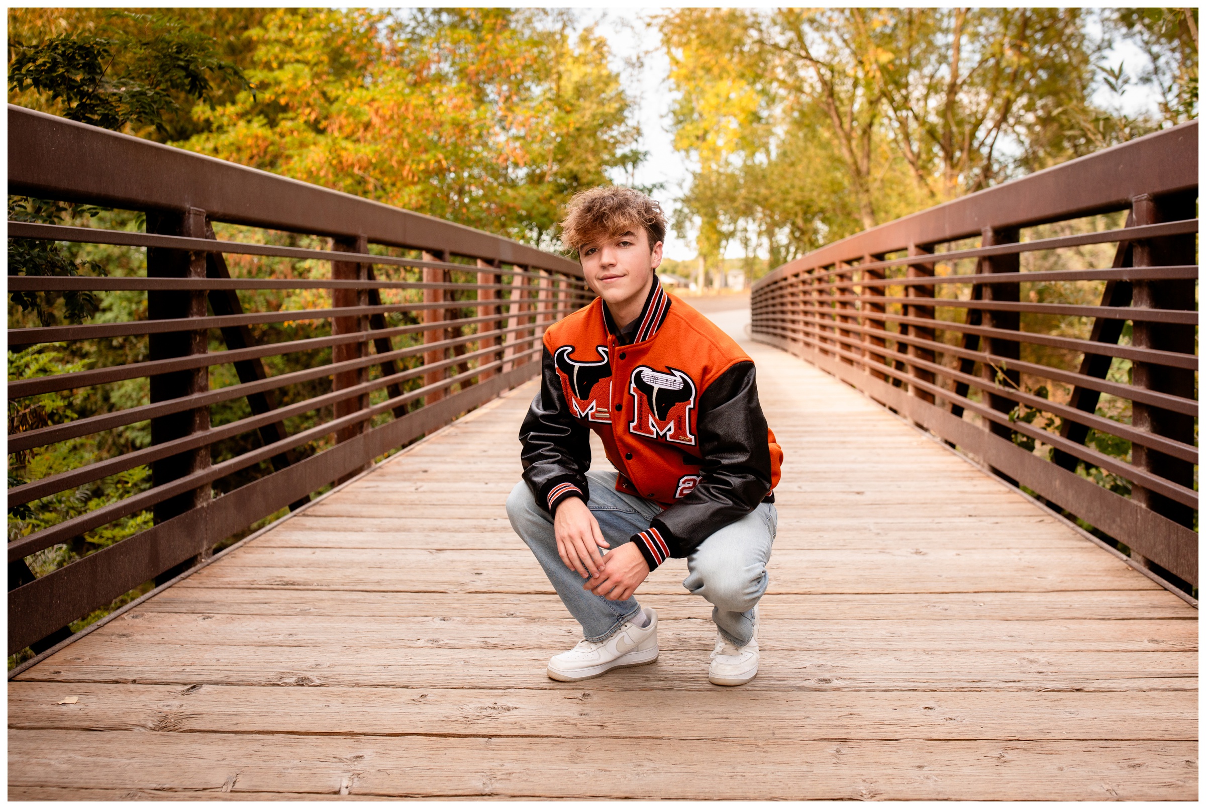 teen squatting on bridge in letterman jacket during Mead High School senior portraits in Colorado